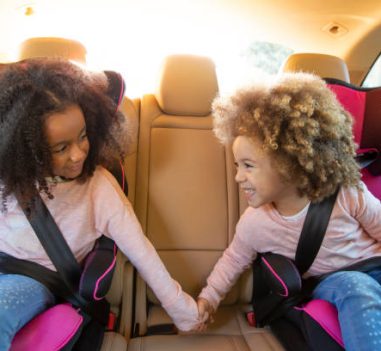 Ethnic kid sister girls in car back seat with safety belts holding hands smiling
